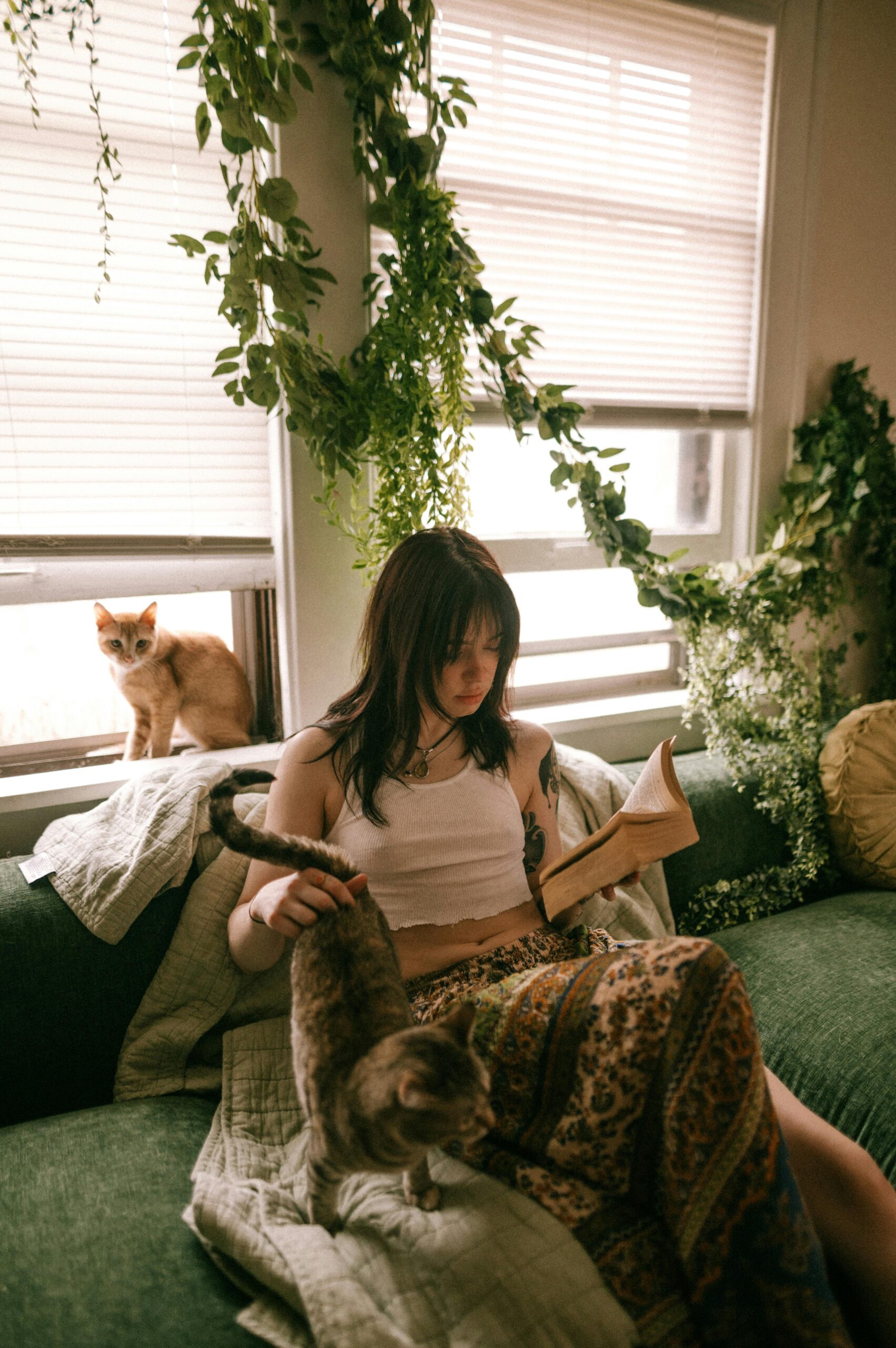 A woman reading on a sofa with cats and lush indoor plants creating a serene atmosphere.