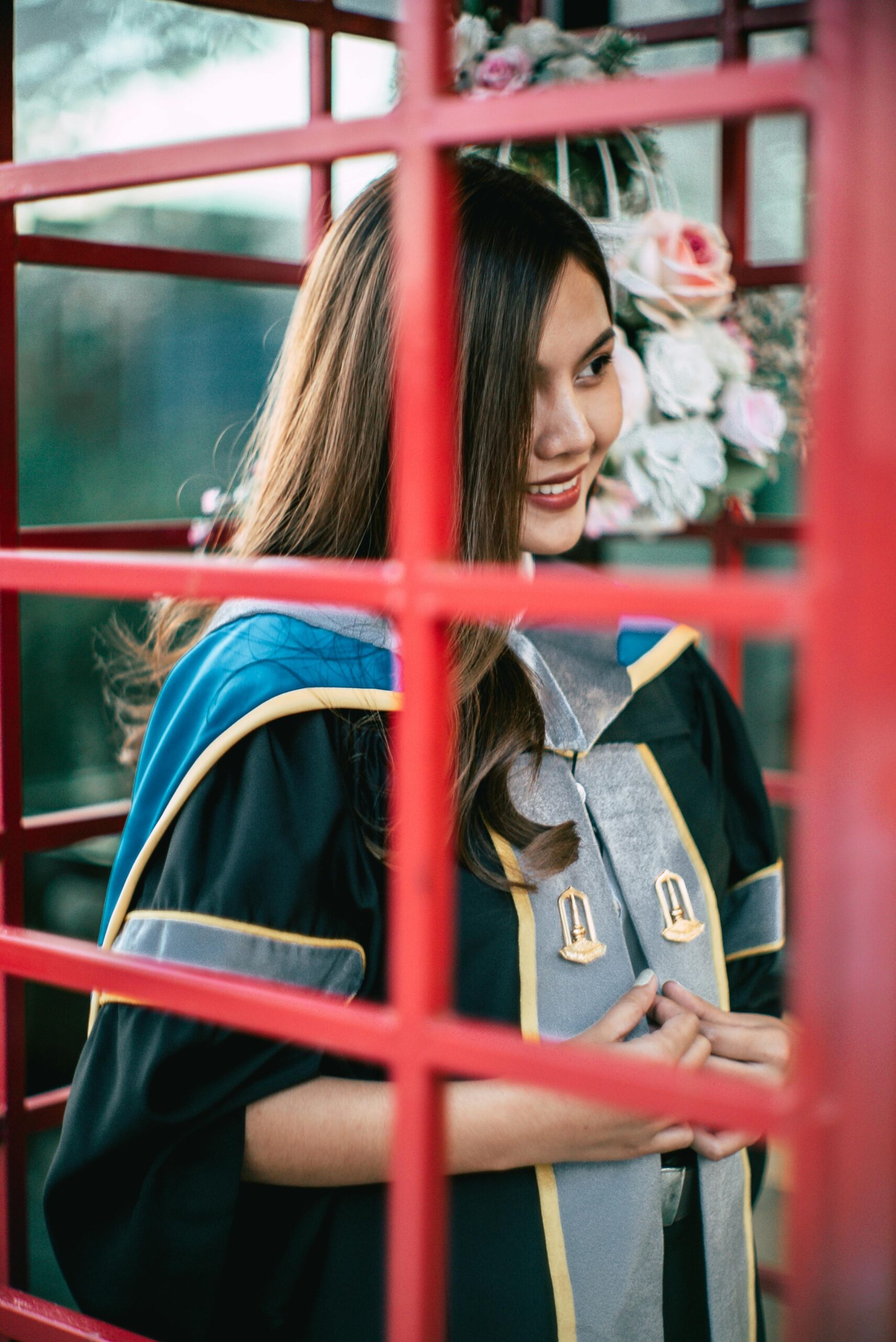 Young woman celebrating graduation in traditional gown inside a red phone booth.
