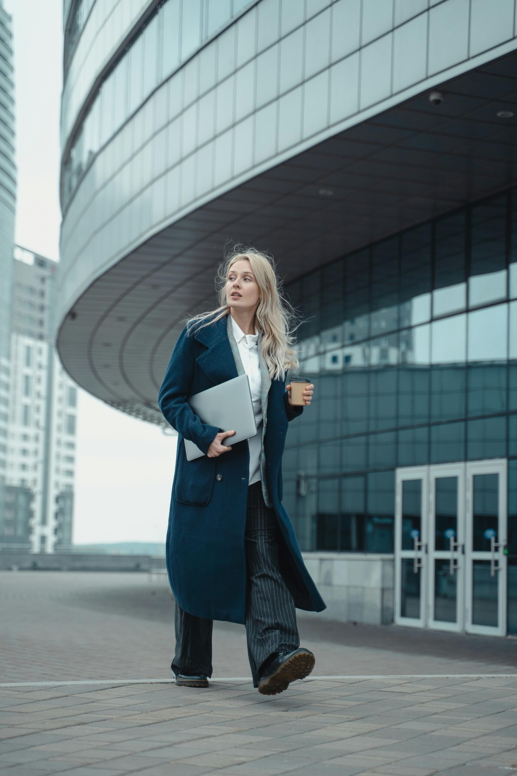 Modern businesswoman with laptop and coffee walking in urban setting.